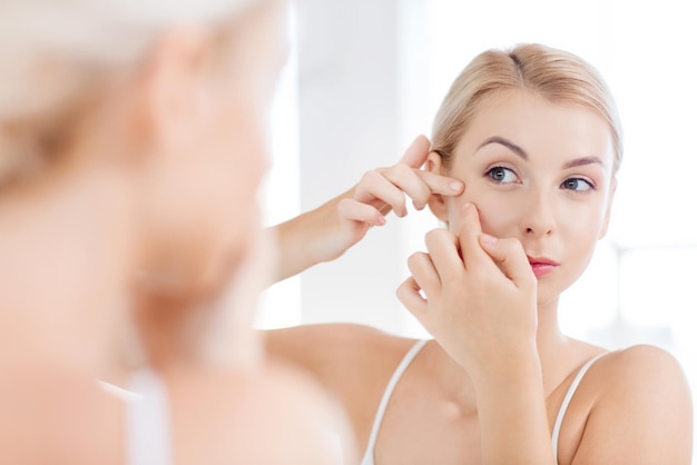 bellezza, igiene, problema della pelle e concetto di persone - giovane donna che guarda allo specchio e schiaccia il brufolo nel bagno di casa