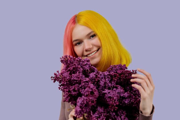 Bellezza, moda, tendenze, acconciature, stile, colore. Ragazza adolescente con capelli tinti di rosa giallo che tiene in mano fiori lilla, con trucco naturale, sorriso con denti bianchi sani, su sfondo viola