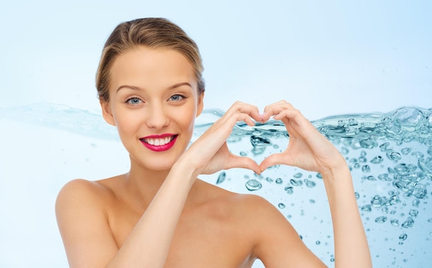 bellezza, persone, amore, giorno di San Valentino e concetto di trucco - giovane donna sorridente con rossetto rosa sulle labbra che mostra il segno della mano a forma di cuore su sfondo di spruzzi d'acqua