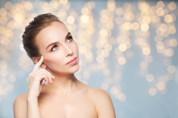 bellezza, persone, vacanze e concetto di cura della pelle - bella giovane donna che tocca il viso su sfondo di luci