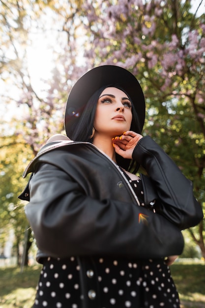 Bellezza soleggiata ritratto donna chic in abito elegante nero in cappello alla moda in giacca di pelle vintage nel parco in giornata di sole luminoso. Splendida bella ragazza bruna e luce solare sulla natura in primavera.