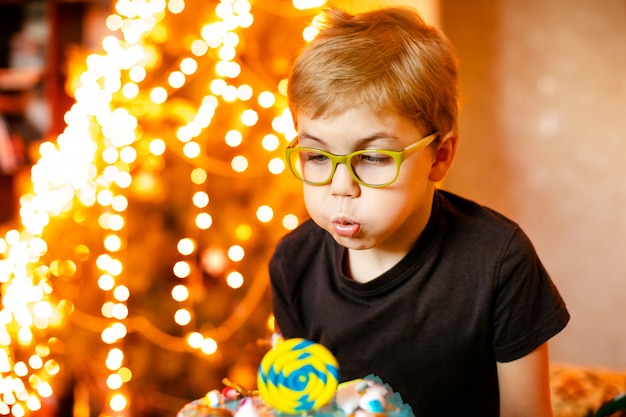 Bellissimo adorabile bambino di sette anni in camicia grigia, festeggia il suo compleanno, soffiando candele sulla torta al forno fatta in casa, coperta