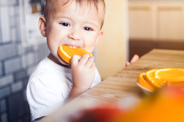 Bellissimo bambino in cucina che mangia avidamente un'arancia, tagliata a spicchi