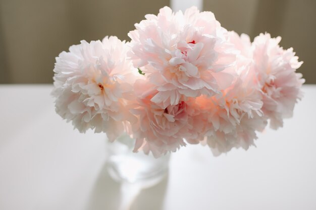 Bellissimo bouquet rosa chiaro di peonie rosa in un vaso di vetro su un tavolo bianco vicino a una finestra