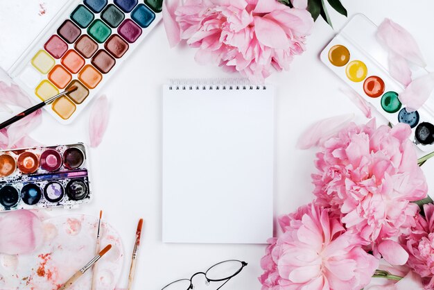 Bellissimo modello femminile flatlay con notebook, articoli di cancelleria, acquerelli e peonie rosa su bianco