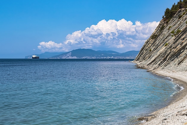 Bellissimo paesaggio estivo, cielo azzurro con nuvole, ripide scogliere con alberi, spiaggia selvaggia di pietra e una vista della città di Novorossijsk all'orizzonte. Russia, Gelendzhik, costa del Mar Nero