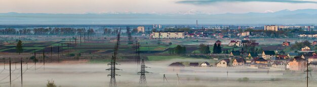 Bellissimo panorama della lunga linea elettrica ad alta tensione elettrica che si estende fino all'orizzonte attraverso campi di primavera e alberi sotto la fitta nebbia mattutina sulla catena montuosa dei Carpazi lontani.