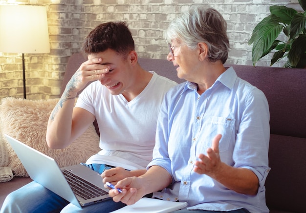 Bellissimo ragazzo seduto sul divano a casa che aiuta la nonna anziana a navigare con il laptop sul web mentre prende appunti Le generazioni più giovani si prendono cura dei parenti più anziani insegnando a usare il computer