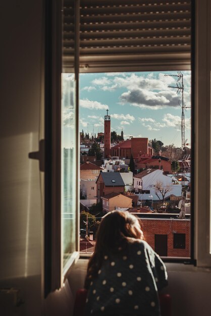 Bellissimo scatto di una bambina con i capelli scuri che guarda fuori dalla finestra in una giornata di sole