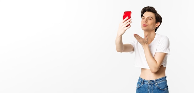 Bellissimo uomo gay in crop top che invia un bacio d'aria alla fotocamera del telefono prendendo selfie o chat video su smartphone in piedi su sfondo bianco