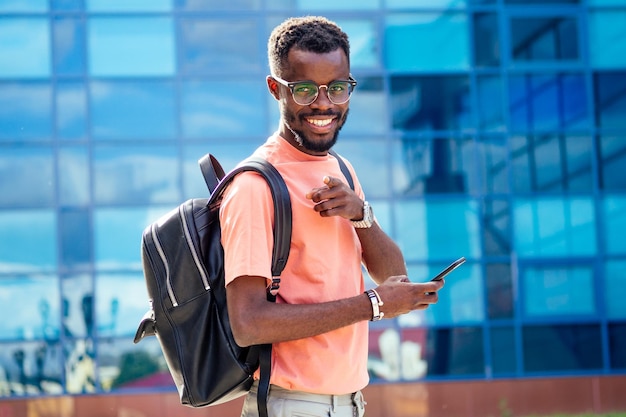 Bello alla moda afroamericani nerd maschio con gli occhiali in abiti eleganti maglietta colorata con uno zaino sulle spalle in piedi sullo sfondo delle finestre blu dell'università