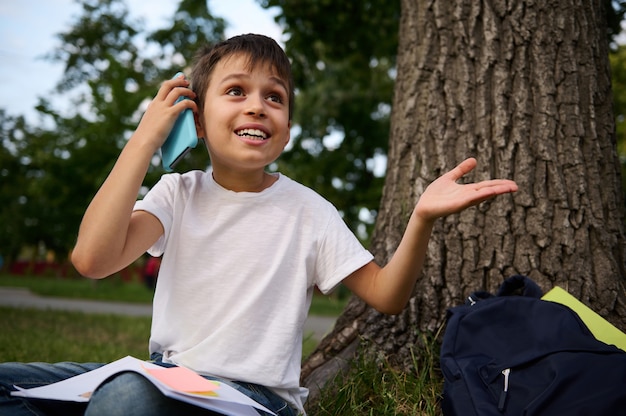 Bello allegro adorabile scolaro che parla al telefono cellulare, carino sorridente che distoglie lo sguardo, gesticolando con le mani, riposando nel parco pubblico dopo il primo giorno di scuola in una bella giornata estiva.