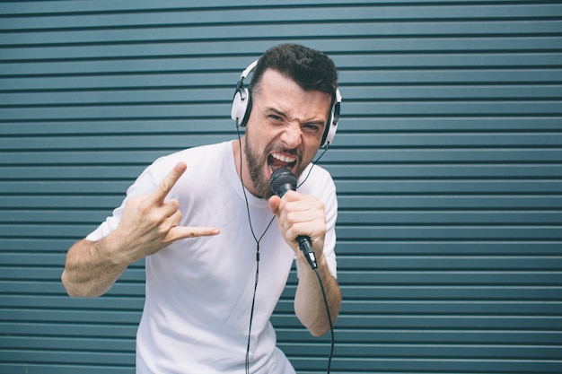 Bello amico è in piedi e in posa sulla macchina fotografica. Sta urlando nel microfono e mostrando il segno cool. L'uomo indossa le cuffie. Lui sta ascoltando la musica. Isolato su strisce