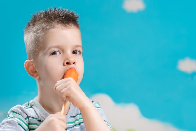 Bello bambino mangia la carota fresca su un bastone. Il ragazzo rosicchia le verdure su uno sfondo blu.
