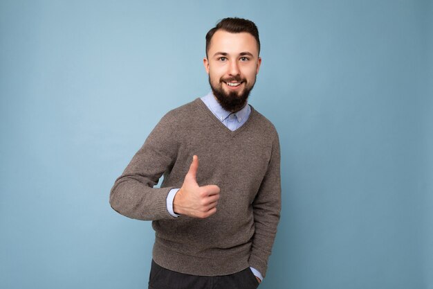 Bello bello brunet barbuto giovane uomo che indossa un maglione grigio e camicia blu isolato su sfondo rosa con spazio vuoto che guarda l'obbiettivo e che mostra i pollici aumenta il gesto.