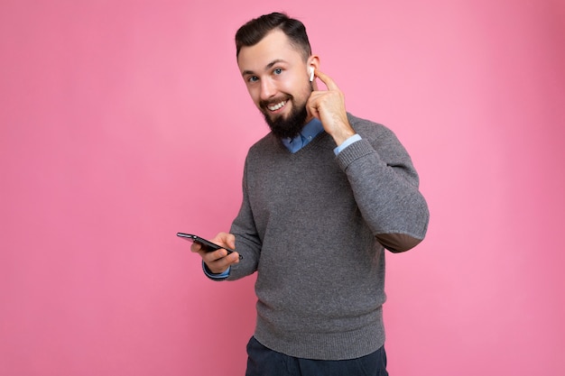 Bello bello brunet barbuto giovane uomo che indossa un maglione grigio e maglietta blu isolato su sfondo rosa con spazio vuoto tenendo in mano e utilizzando il telefono cellulare comunicare online guardando c