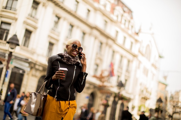 Bello caffè alla moda della tenuta della donna afroamericana e facendo uso di uno Smart Phone