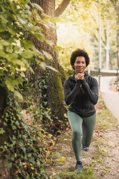 Bello corridore femminile sorridente dell'afroamericano che fa gli esercizi di affondi al mattino in natura.