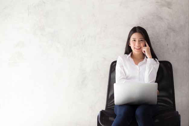 Bello del sorriso asiatico della giovane donna del ritratto facendo uso del computer portatile nel luogo di lavoro