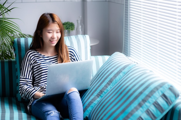 Bello della donna asiatica del ritratto che per mezzo del computer portatile per svago sul sofà