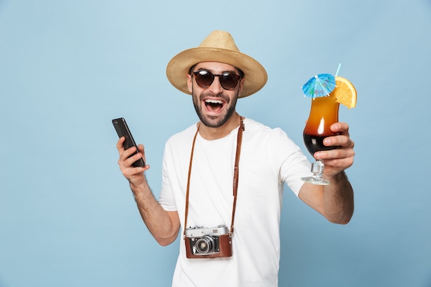 bello eccitato giovane turista in posa con la fotocamera utilizzando il telefono cellulare bevendo cocktail isolato sul muro blu.