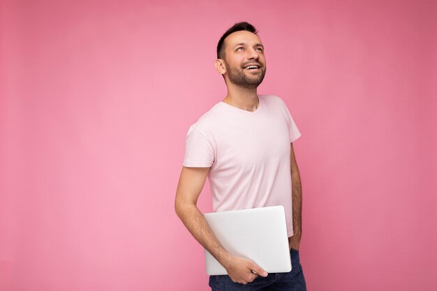 Bello felice giovane uomo con la barba lunga che tiene il computer portatile che osserva in su in maglietta sulla superficie rosa isolata