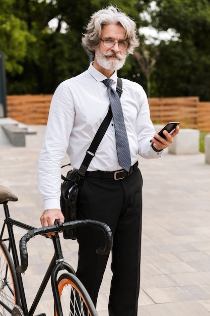 Bello fiducioso uomo d'affari barbuto che indossa tuta, porta una valigetta camminando all'aperto con la bicicletta, usando il telefono cellulare