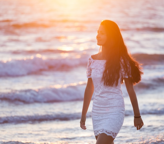 Bello giovane adolescente con un vestito bianco sulla spiaggia al tramonto