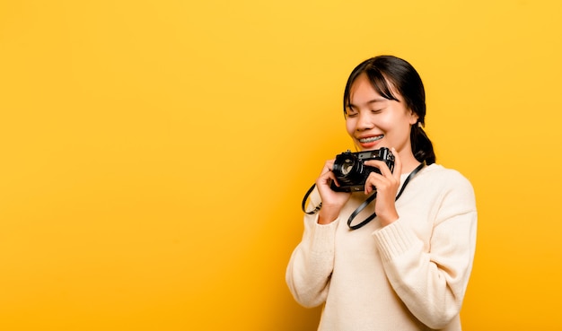 Bello giovane studio turistico asiatico girato con la macchina fotografica a disposizione isolata su fondo giallo. concetto di fotografia