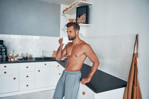 Bello giovane uomo a torso nudo che beve tè caldo mentre si trova in cucina a casa