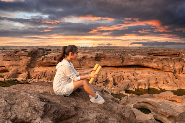 Bello giovane viaggiatore asiatico della donna che si siede e che esamina la mappa di carta sulla scogliera di roccia nel grand canyon della Tailandia al tramonto