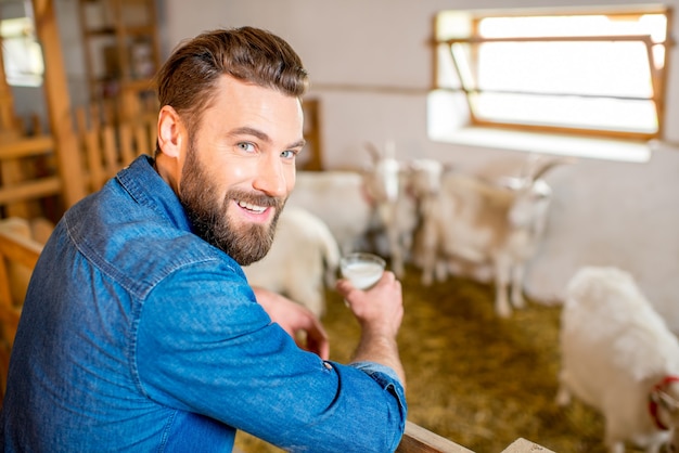 Bello l'agricoltore che assaggia il latte fresco in piedi nella stalla con capre sullo sfondo. Produzione e allevamento di latte naturale