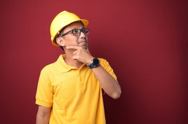 Bello lavoratore asiatico che indossa il casco che pensa con la mano sul mento su sfondo giallo.