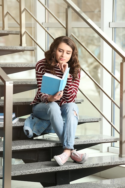 Bello libro di lettura della giovane donna mentre sedendosi sulle scale vicino alla finestra