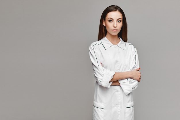 Bello modello femminile in uniforme medica che posa al fondo grigio. Giovane donna in camice medico, copia spazio per la tua pubblicità. Concetto di assistenza sanitaria