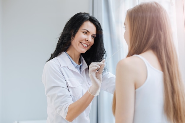 Bello pediatra positivo qualificato che lavora nell'ospedale e che ha appuntamento con il paziente mentre utilizza la lama medica per l'esame della gola