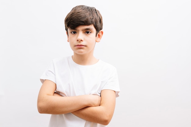 Bello ragazzo del bambino che porta la maglietta casuale grigia che sta sopra il fronte bianco isolato del fondo serio con le braccia incrociate che esamina la macchina fotografica.