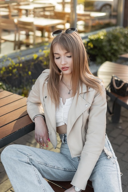 Bello ritratto femminile alla moda di una ragazza piuttosto giovane hipster in un vestito di moda di strada con giacca rock in pelle e jeans blu seduto su una panchina fuori da un caffè in città