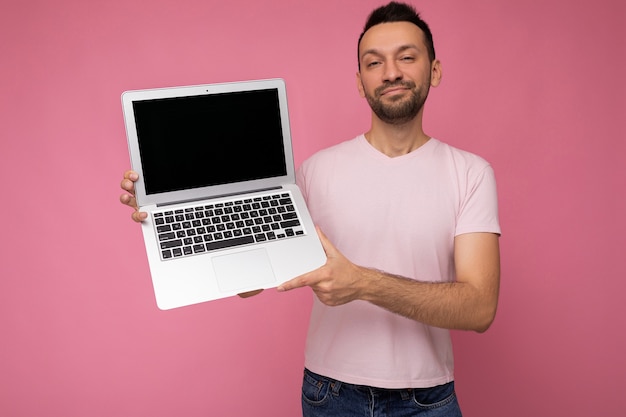 Bello sorridente brunet uomo che tiene il computer portatile in t-shirt sul muro rosa.