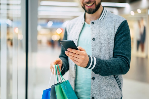 Bello sorridente giovane uomo barbuto elegante con borse della spesa sta utilizzando smart phone mentre si cammina nel centro commerciale