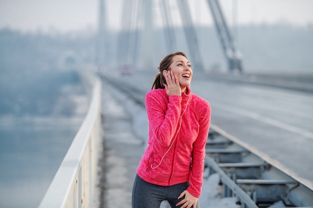 Bello sorridere castana caucasico nella musica di riposo e d'ascolto degli abiti sportivi mentre stando sul ponte. Orario invernale. Fitness all'aperto.