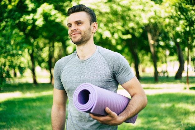 Bello sportivo, magro e forte sportivo o istruttore di yoga con tappetino yoga in mano sullo sfondo del parco cittadino