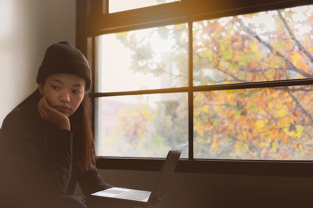 Bello studente asiatico della donna che pensa e che lavora con il computer portatile nella sala.