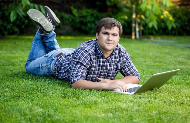 Bello studente sorridente sdraiato sull'erba con il computer portatile