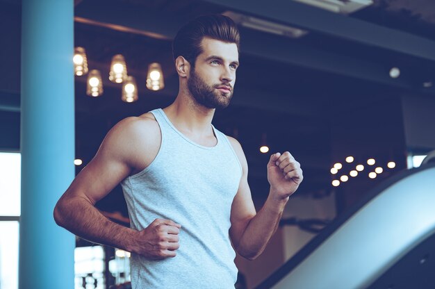 Bello sul tapis roulant. Inquadratura dal basso del giovane bell'uomo in abbigliamento sportivo che corre sul tapis roulant e guarda lontano in palestra