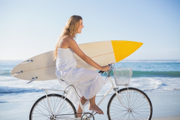 Bello surfista in prendisole sulla bici che tiene tavola da surf in spiaggia