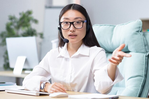 Bello tecnico di supporto della banca femminile asiatica che esamina la macchina fotografica e parla durante la videochiamata utilizzando