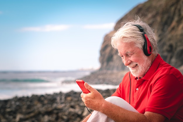 Bello uomo anziano in musica d'ascolto rossa dalle cuffie che si siedono sulla spiaggia uomo felice anziano in mare che tiene smartphone che gode del tempo libero e della pensione