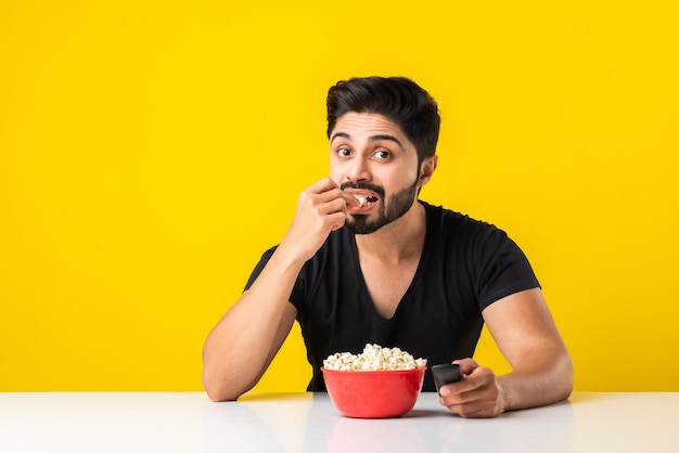 Bello uomo barbuto Indin che mangia popcorn dalla ciotola rossa mentre è seduto a tavola su sfondo giallo