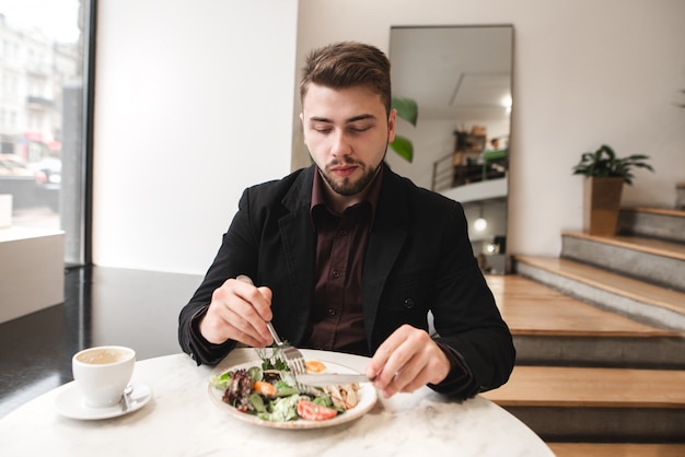 Bello uomo con la barba e un vestito seduto a un tavolo in un accogliente caffè e mangiare insalata e bere caffè